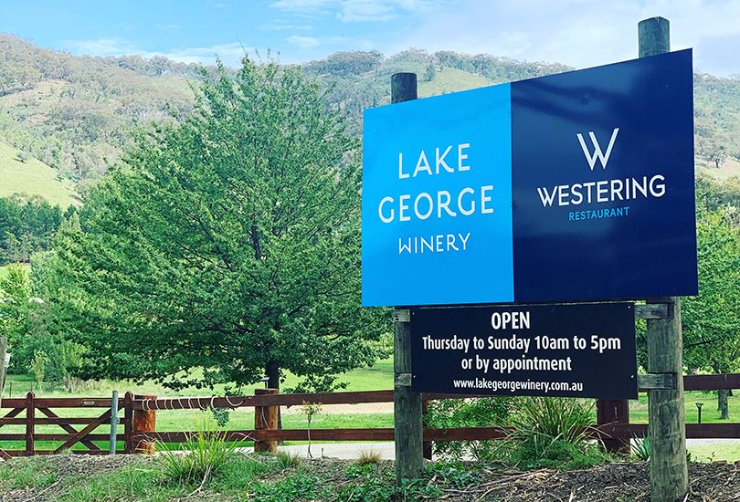 Sign at entrance to Lake George Winery, with trees and hills in background.