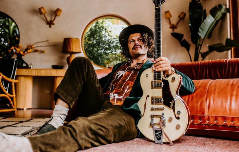 Ash Grunwald leaning against a couch with guitar in hand
