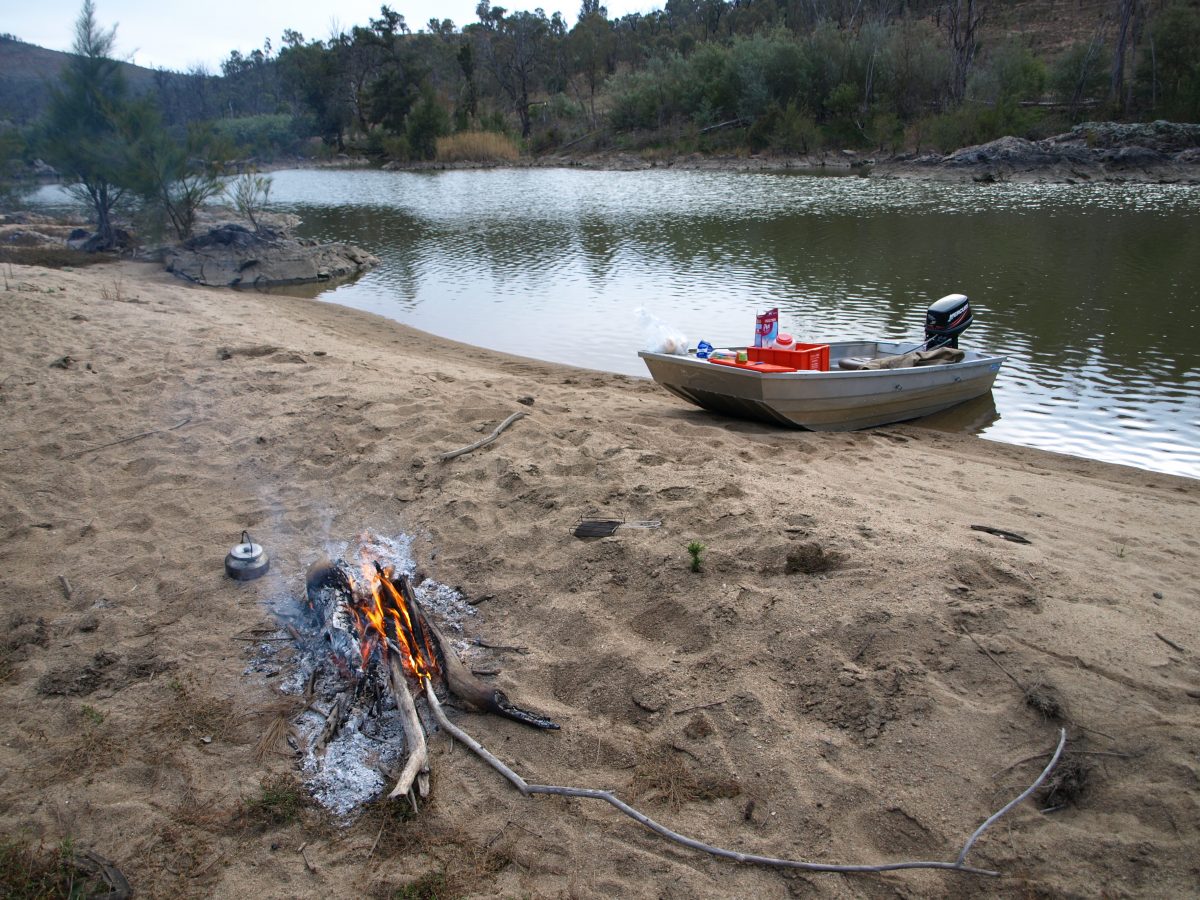 Campfire on the foreshore of the Murrumbidgee river, nearby a tinny boat is beached.