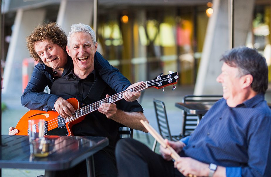 Two men sitting at a cafe table with Leo Sayer leaning over one of them and playing the guitar