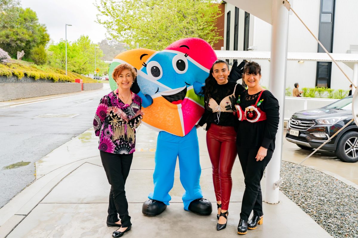 Three women standing with a colourful heart mascot, wearing embellished bras on the outside of their shirts as part of the 'Bra Walk' fundraiser for Can Give Day.