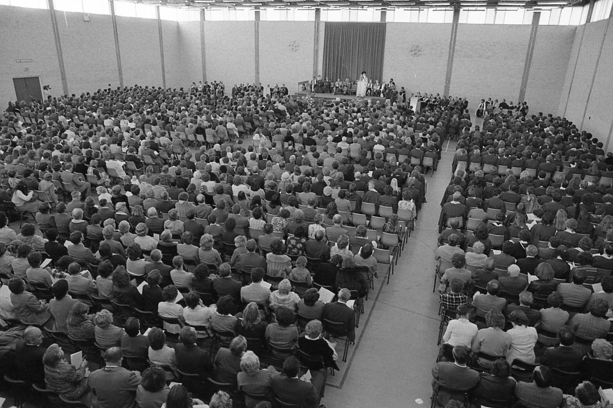 A black-and-white aerial view of a graduation ceremony at the University of Canberra in the 1970s. Rows of graduates face a stage on which a speaker stands.
