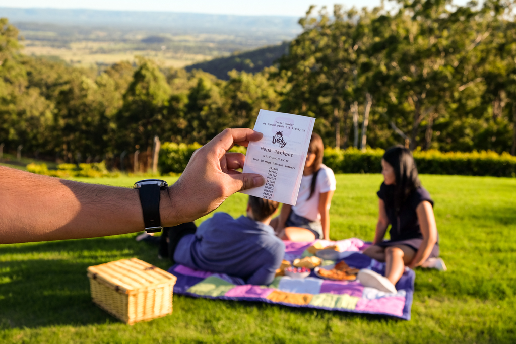 A hand holding out a lottery ticket with a group of people having a picnic on the side of a hill in the background