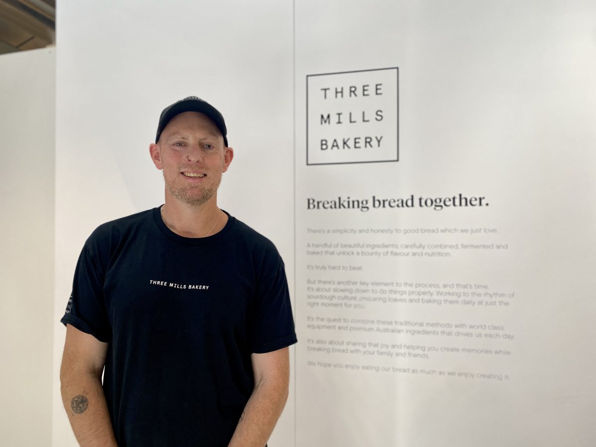 Ben wears black Three Mills Bakery shirt in front of wall with text "Three Mills Bakery, baking bread together"