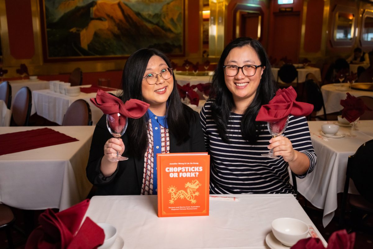 Jennifer and Lin are both Asian women wearing glasses and smiling, while holding wine glasses stuffed with a red napkin. A copy of the book Chopsticks or Fork? sits on the table.