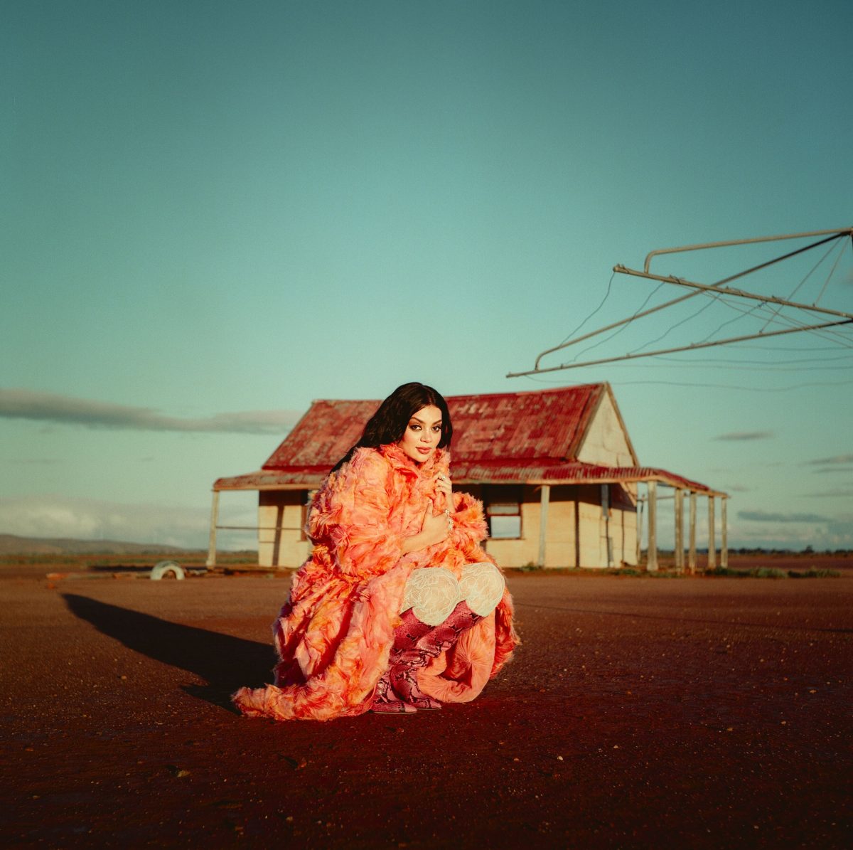 A woman in a flowing red dress crouched near an old house in the country 