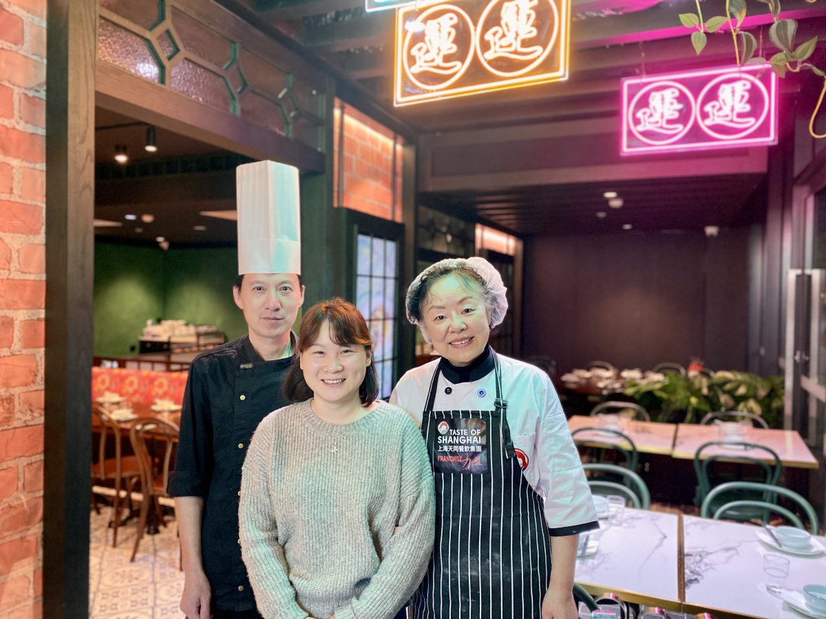 A man in tall chef's hat stands with two women (one with apron and hairnet) in a restaurant with colourful neon signs.